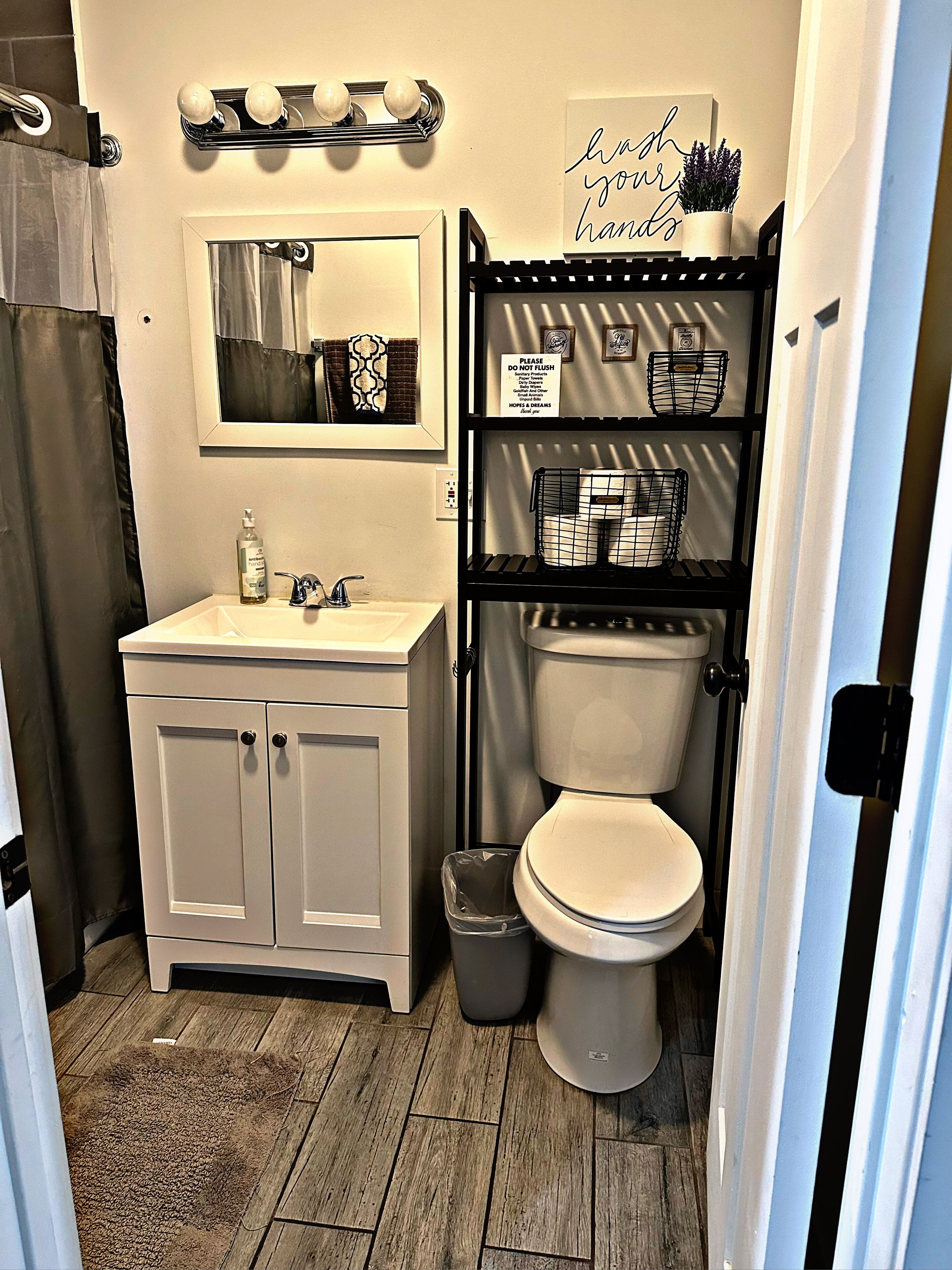 A bathroom with a white sink, mirror, and toilet. There is a shelf with toiletries and a sign above the toilet.