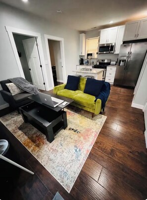 A modern living room with a sofa, coffee table, and a laptop. A kitchen with appliances is visible in the background.