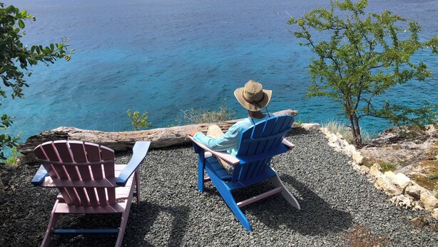 A person sitting on a blue chair with a hat, overlooking a body of water.