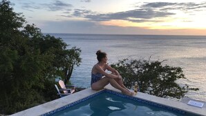 A person sitting by a pool with a view of the sea at sunset.