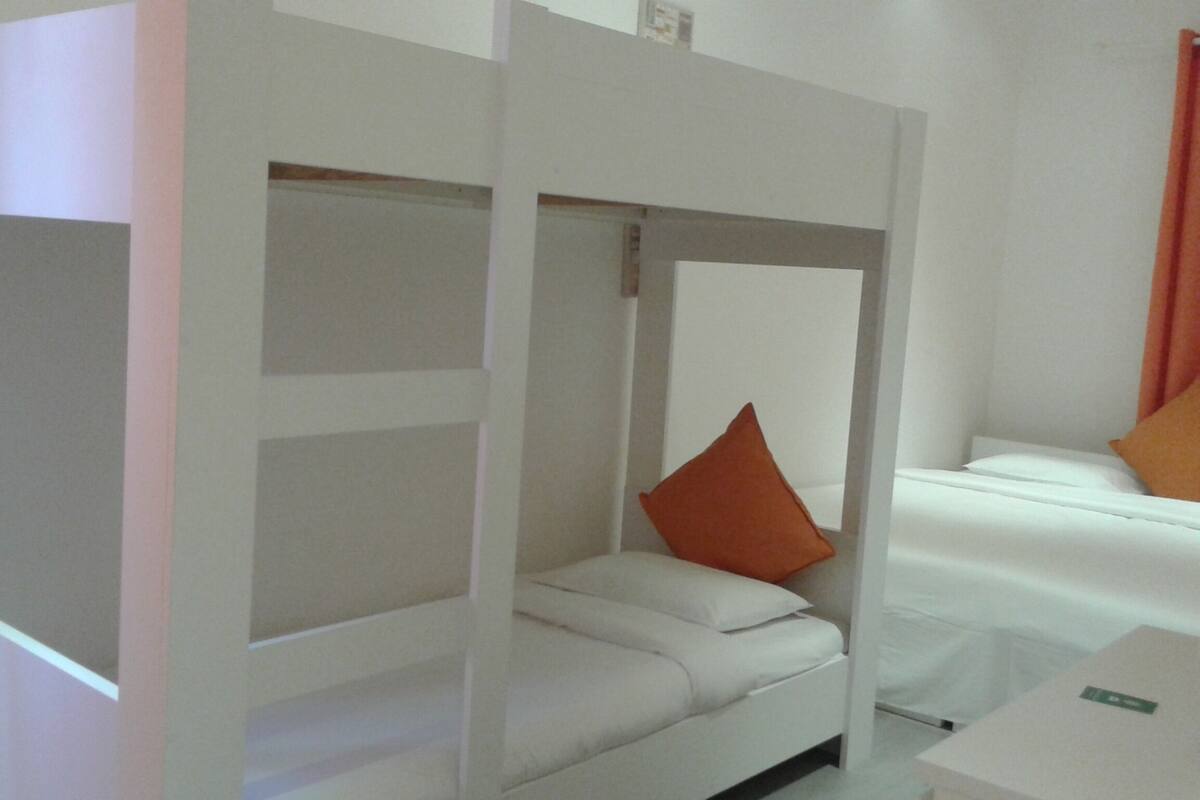 Une chambre avec un lit superposé et un lit simple. Il y a un mur blanc et une fenêtre avec des rideaux.