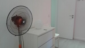Une pièce avec un bureau blanc, un ventilateur sur pied, une chaise blanche et une porte fermée.
