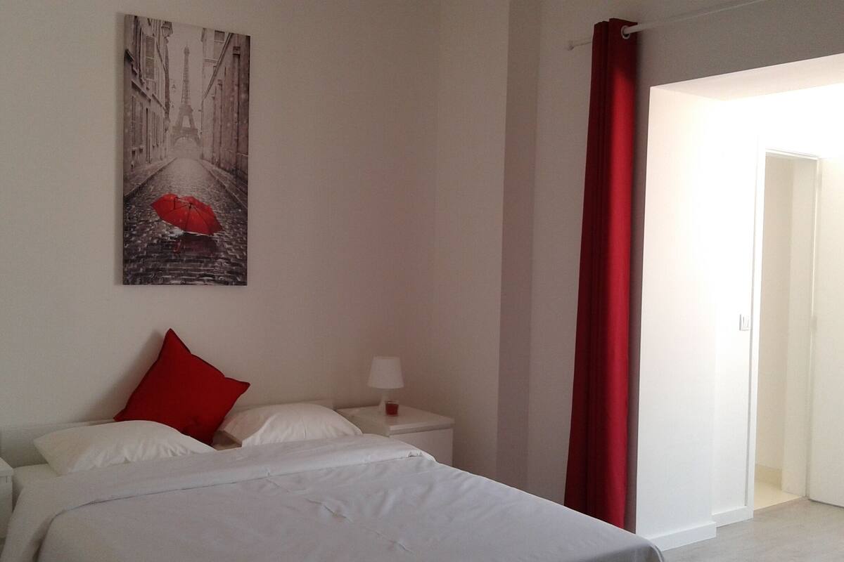 Une chambre d’hôtel avec un lit, une table de chevet, une lampe, un rideau rouge et un tableau représentant la Tour Eiffel.
