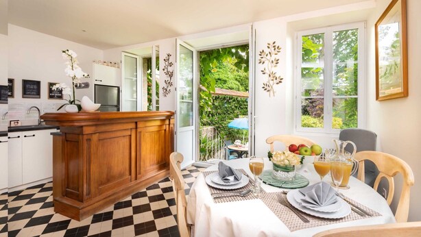 A dining area with a checkered floor, a wooden dining table set with plates, glasses, and a fruit bowl. A kitchen with white cabinets and a black and white checkered floor is visible in the background.