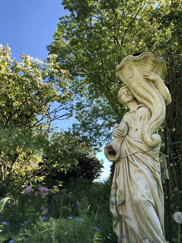 A statue of an angel with outstretched wings, set in a garden with lush greenery and flowering plants.