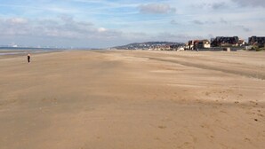 A wide sandy beach with a few people walking, buildings in the background, and a clear sky.