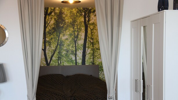 A bedroom with a bed, curtains, a wardrobe, and a wall with a forest mural. - Apartment am Kurpark - beautiful, quiet and right in the middle -  (St. Andreasberg)