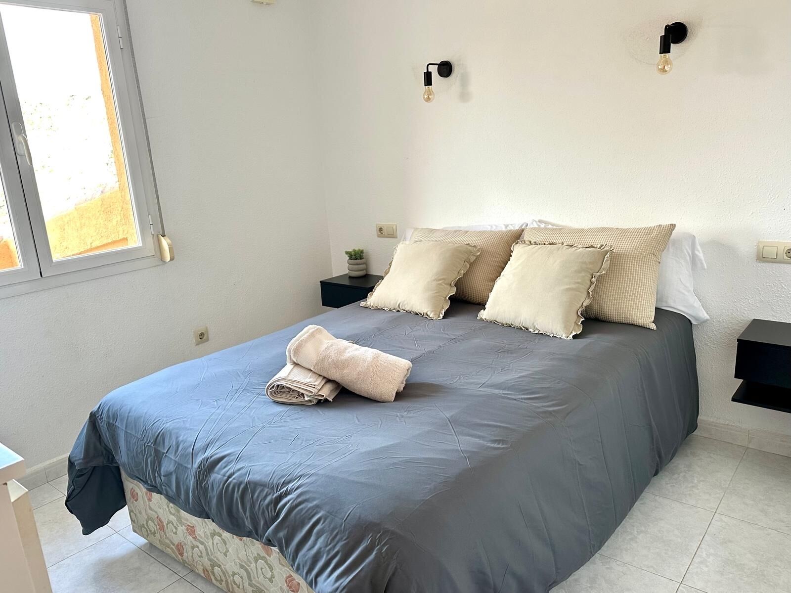 A neatly made bed with a blue and grey duvet, two beige pillows, and a folded towel on the bed. There are two bedside tables, one with a small plant and the other with a lamp.