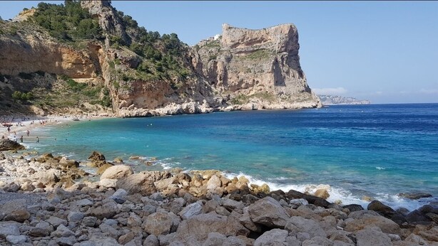 A rocky beach with clear blue water, a large cliff, and a castle-like structure. - Magnificent quiet apartment close to everything with swimming pool. (Benitachell)