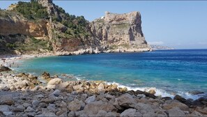 Ein felsiger Strand mit klarem, blauem Wasser, eine hohe Klippe und ein schlossähnliches Gebäude.