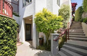 A residential building with a staircase leading to a door, surrounded by greenery and a red railing.