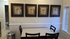 Un coin repas avec une table blanche et quatre chaises, des œuvres d’art encadrées au mur et une fenêtre sur le côté droit.