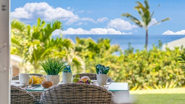 Ein Tisch mit Tassen, einer Obstschale und einer Pflanze, mit Blick auf eine tropische Landschaft mit Palmen und dem Meer.