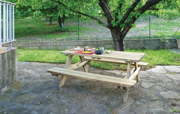 A picnic table with benches and a tablecloth, surrounded by a stone patio and a grassy area with trees. - Beautiful home in Le Poujol sur Orb (Le Poujol sur Orb)