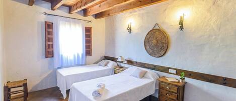 A hotel room with two single beds, wooden headboards, a small wooden table, a nightstand, a wall-mounted lamp, and a woven wall decoration.