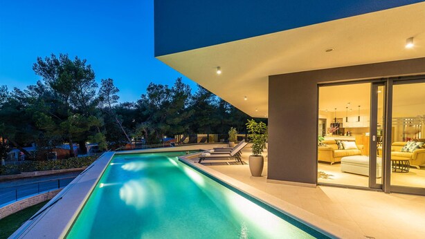 A modern outdoor pool area with lounge chairs, a well-lit interior with a sofa and dining area, and a view of trees and a building in the background. - Cozy home in Biograd na moru with sauna (Biograd na moru)
