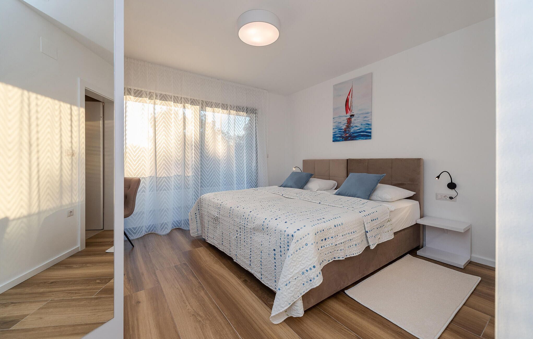 A bedroom with a wooden floor, a bed with a white quilt, a nightstand, a lamp, and a painting of a sailboat.
