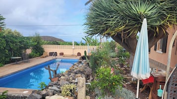 Una piscina con cubierta de madera, rodeada de rocas y plantas, y un patio con mesa y sillas.