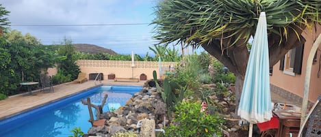 Una piscina con cubierta de madera, rodeada de rocas y plantas, y un patio con mesa y sillas.