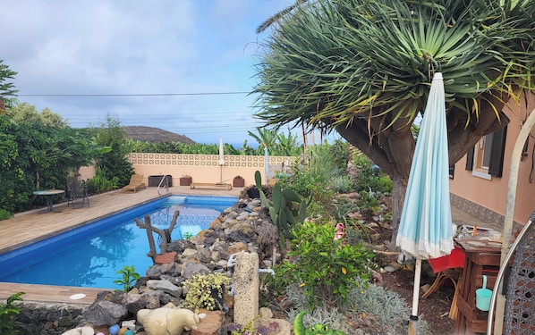 Una piscina con cubierta de madera, rodeada de rocas y plantas, y un patio con mesa y sillas.