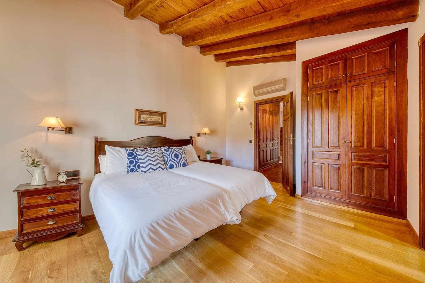 A bedroom with a wooden ceiling, a large bed with patterned pillows, a nightstand with a lamp, and a wardrobe.