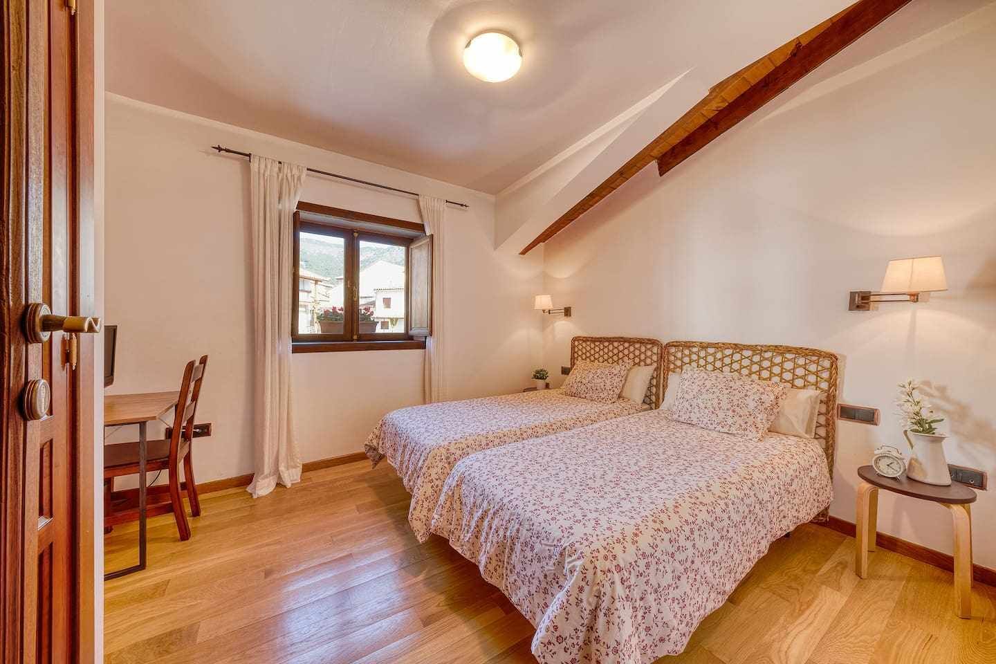 Un dormitorio con dos camas, techo de madera, ventana con cortinas, mesita de noche con lámpara y una silla.