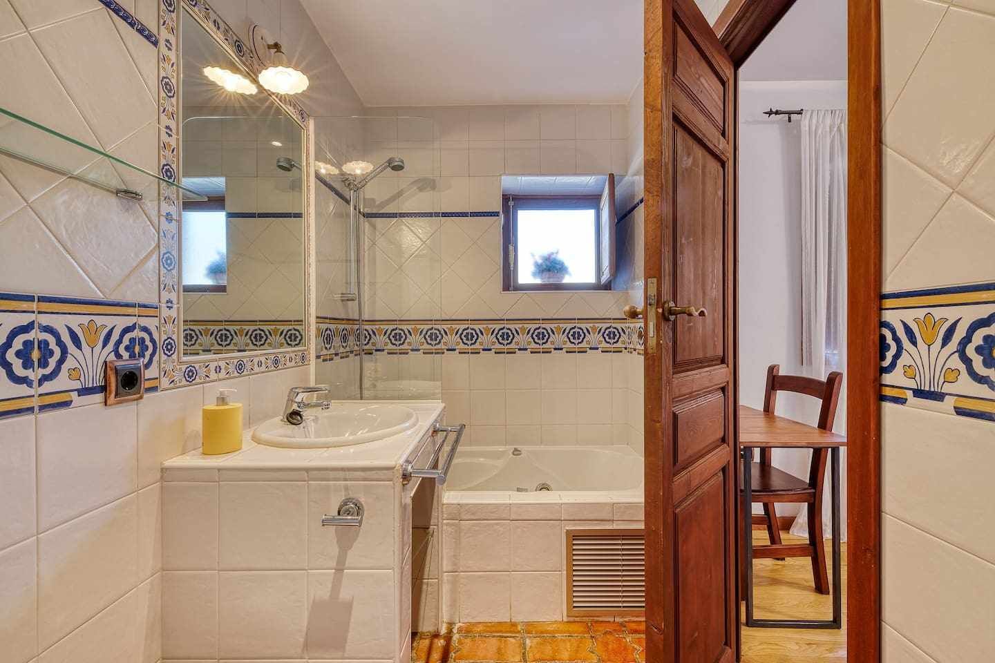 A bathroom with a sink, mirror, and shower. There is a small window above the mirror and a door leading to another room.