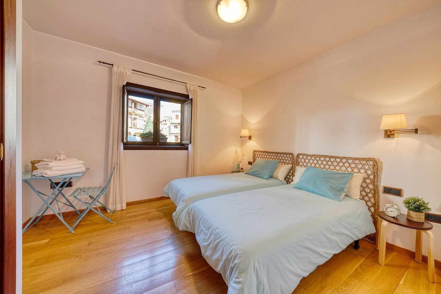 A bedroom with two beds, a window with curtains, a small table with towels, and a nightstand with a lamp.