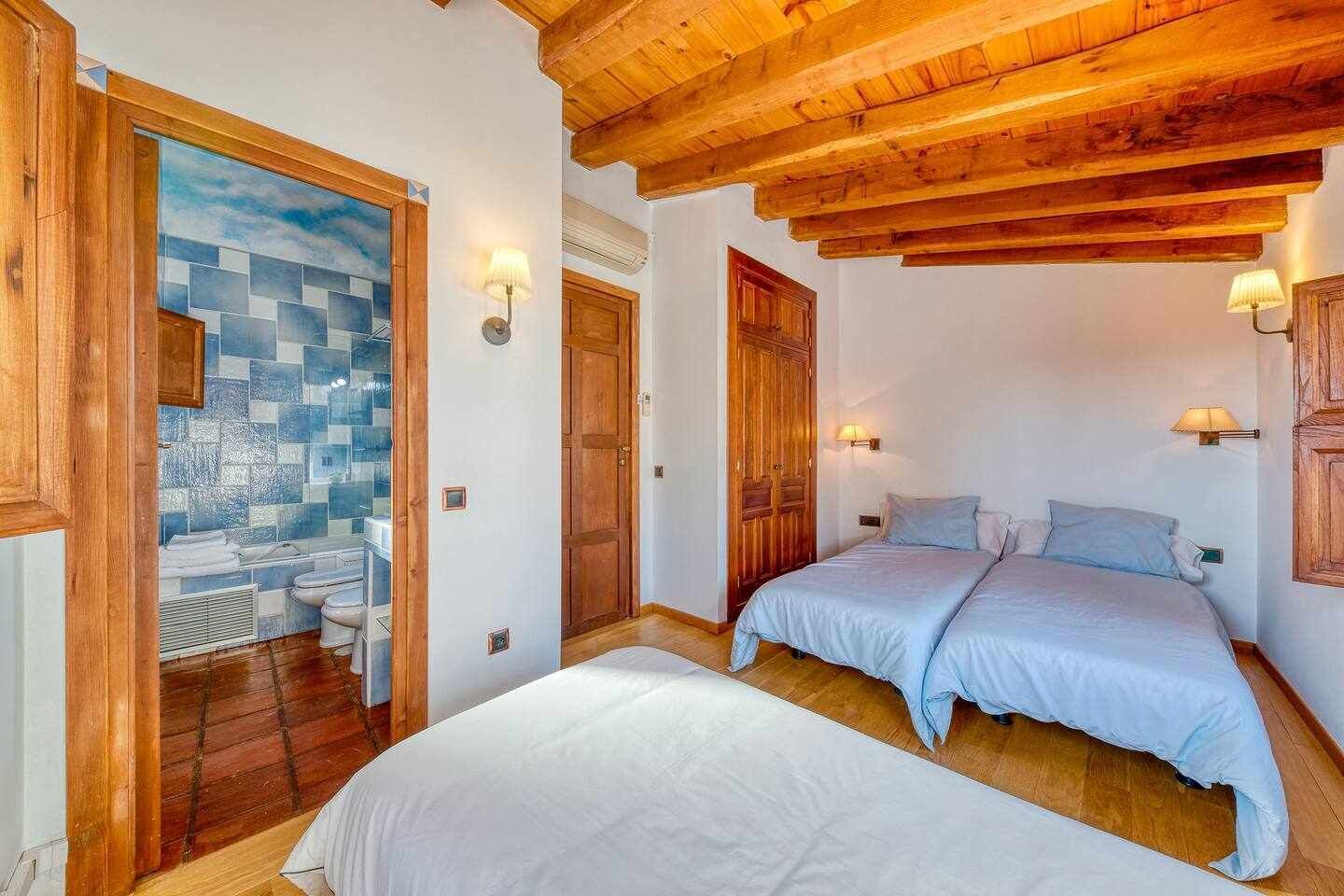 A bedroom with two beds, wooden ceiling beams, and a bathroom with a mosaic tile wall.