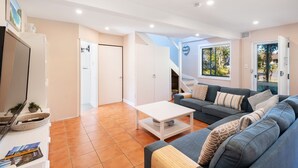 Een moderne woonkamer met een bank, salontafel en een televisie. Er is een deur die leidt naar een andere kamer en een raam met uitzicht.