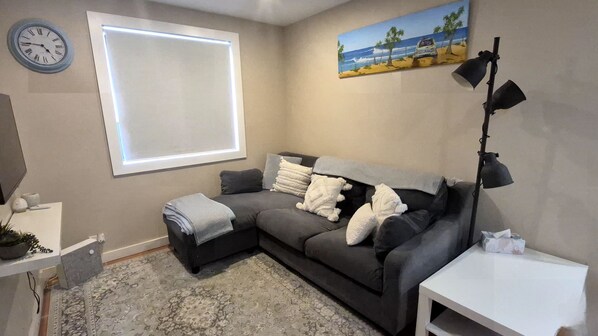 A living room with a grey sectional sofa, a white side table, a floor lamp, a painting of a beach scene, and a wall clock. - Beecroft Street Retreat by Book Jervis Bay - Free Wifi, Linen & Pet Friendly (Huskisson)