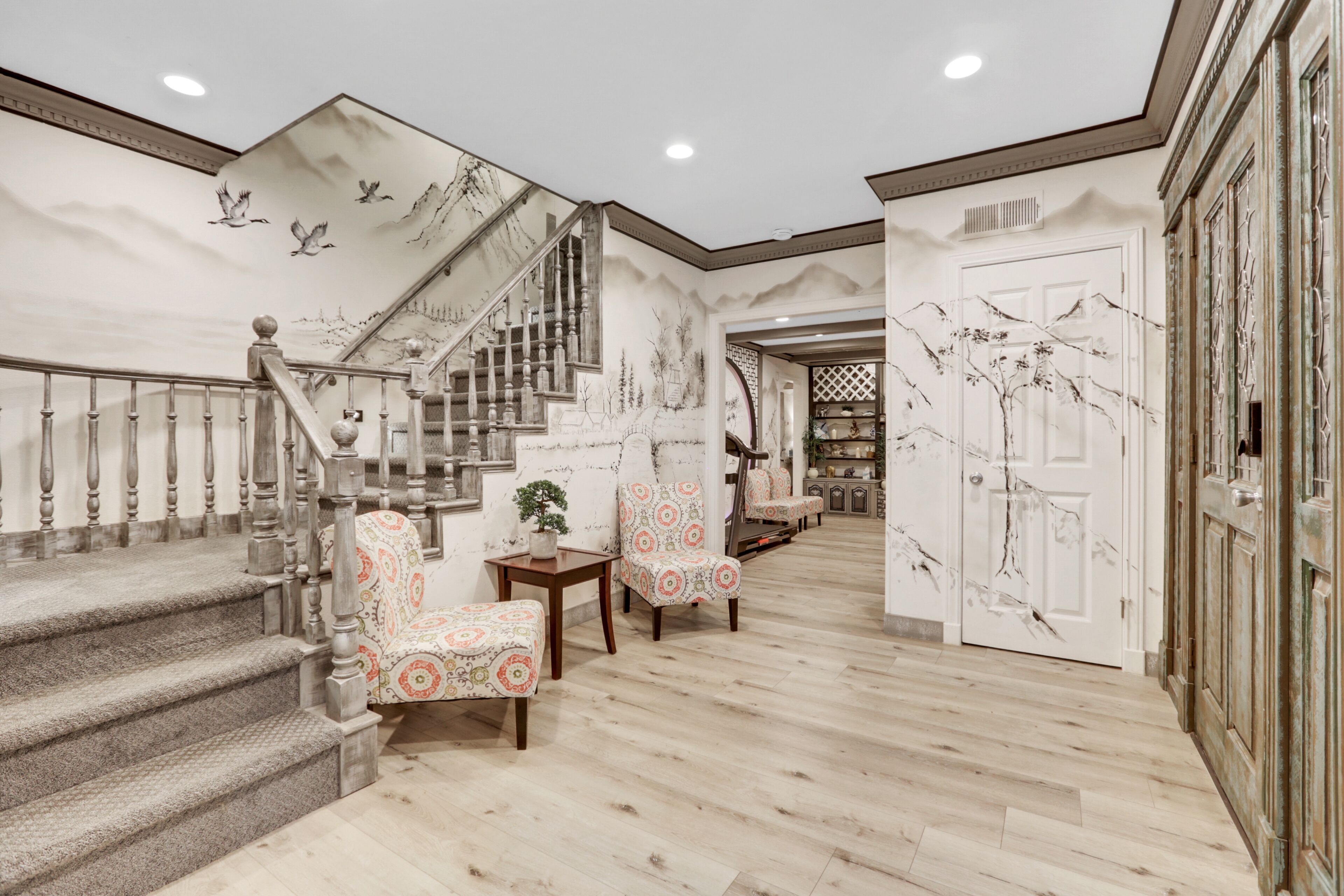 Un hall d’entrée spacieux avec un grand escalier, un coin salon doté de fauteuils à motifs et un mur décoratif orné d’une fresque représentant un arbre.