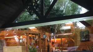 A wooden structure with a glass window, red geraniums in pots, and a view of a rustic interior with wooden beams and a clock.