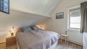 Quarto com teto inclinado, cama com manta estampada, mesa de cabeceira com abajur, um banquinho pequeno, radiador e janela com persianas.