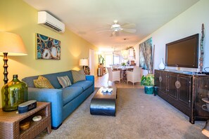 A living room with a blue sofa, a coffee table, a TV on a cabinet, and a dining area in the background.