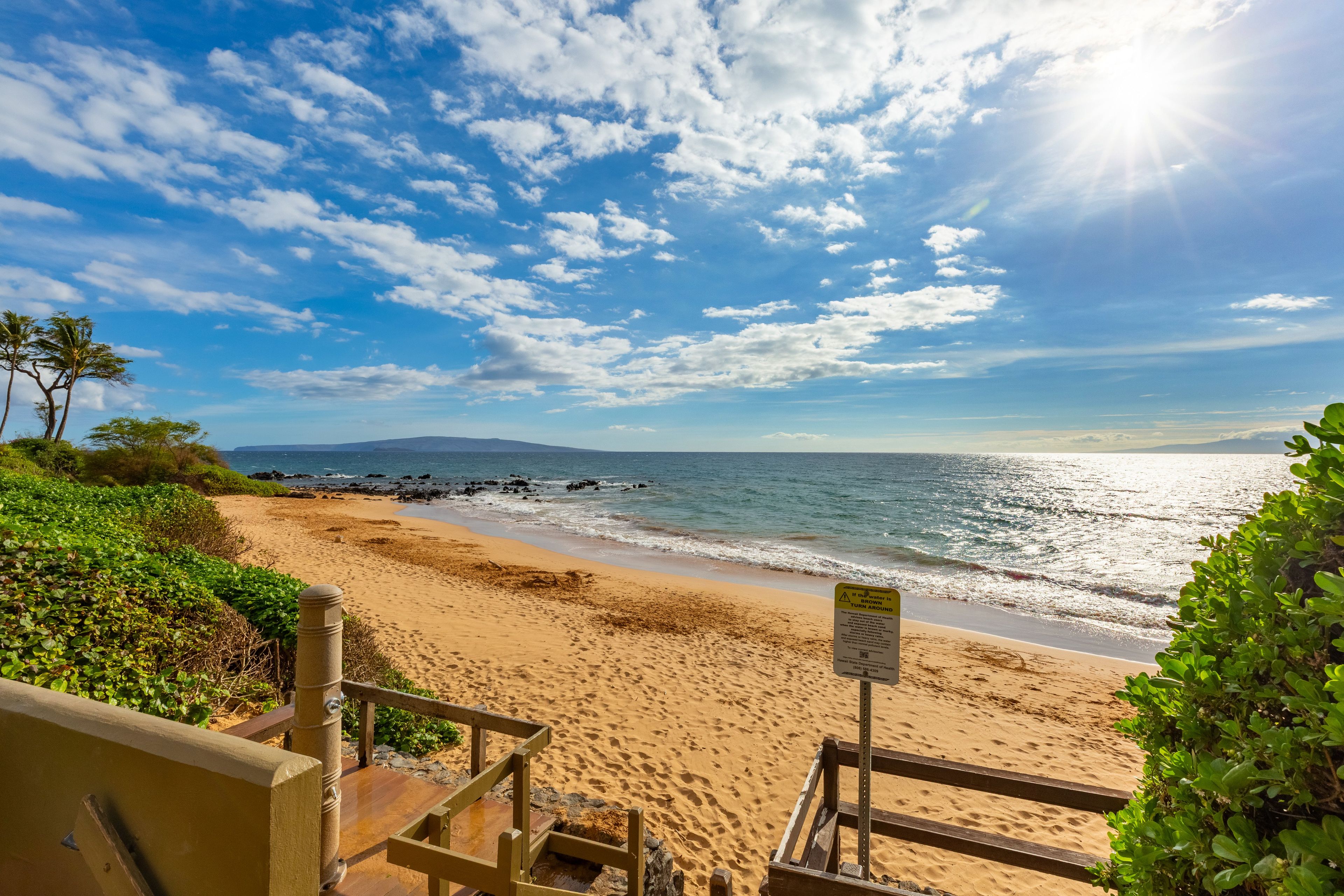 A beach with a wooden staircase leading down to the shore, a sign warning about the water, and a clear view of the ocean and sky.