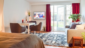 Et hotelværelse med seng, sofa, spisebord med stole, fjernsyn og et stort vindue med gardiner.