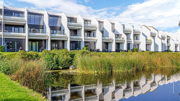 En moderne bygning med flere etager, altaner og en spejlen­de vandflade foran, under en klar himmel.