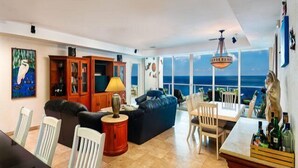 A spacious living room with a large window offering a view of the ocean, a dining area with a table and chairs, and a seating area with a sofa and chairs. - Amazing Ocean Views (Cozumel)
