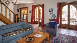 Een woonkamer met een blauwe bank, een houten salontafel en een kamerkleed met een patroon. Er is een trap met houten leuningen, een deur naar buiten en een schilderij aan de muur.