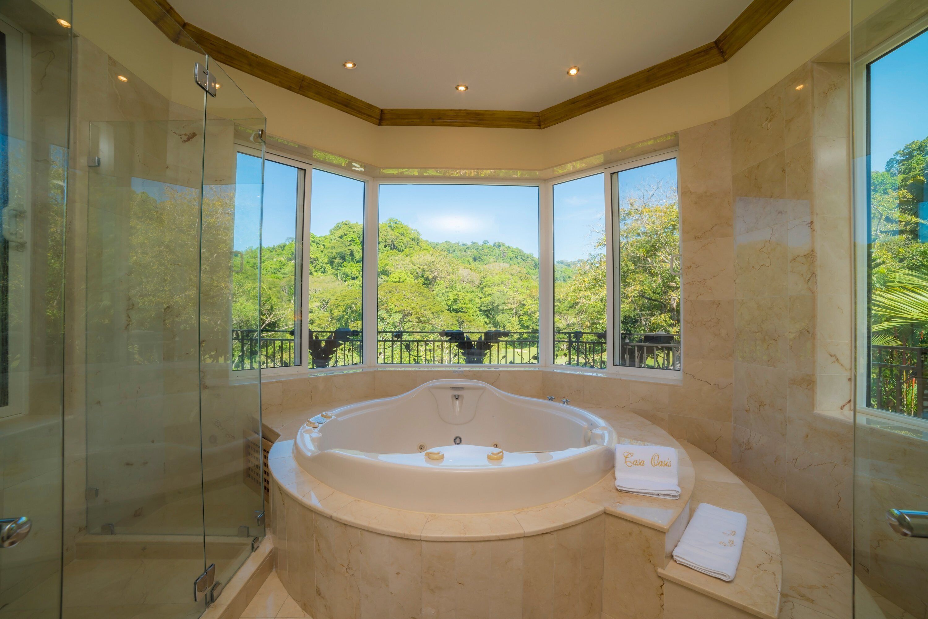 Ein Badezimmer mit einer großen Badewanne, einer Duschkabine aus Glas und Blick auf üppiges Grün durch große Fenster.