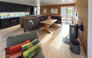Une cuisine moderne dotée d’un four encastré, un coin repas avec une table et des chaises en bois, et un coin salon confortable avec un canapé et un coussin coloré.