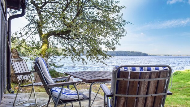 Una terraza de madera con dos sillas y una mesa, con vista a un lago y una orilla distante.