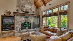 A cozy living room with a stone fireplace, leather sofa, and large windows offering a view of the outdoors. - Holly Hideaway - TLUXP (Tahoe City)