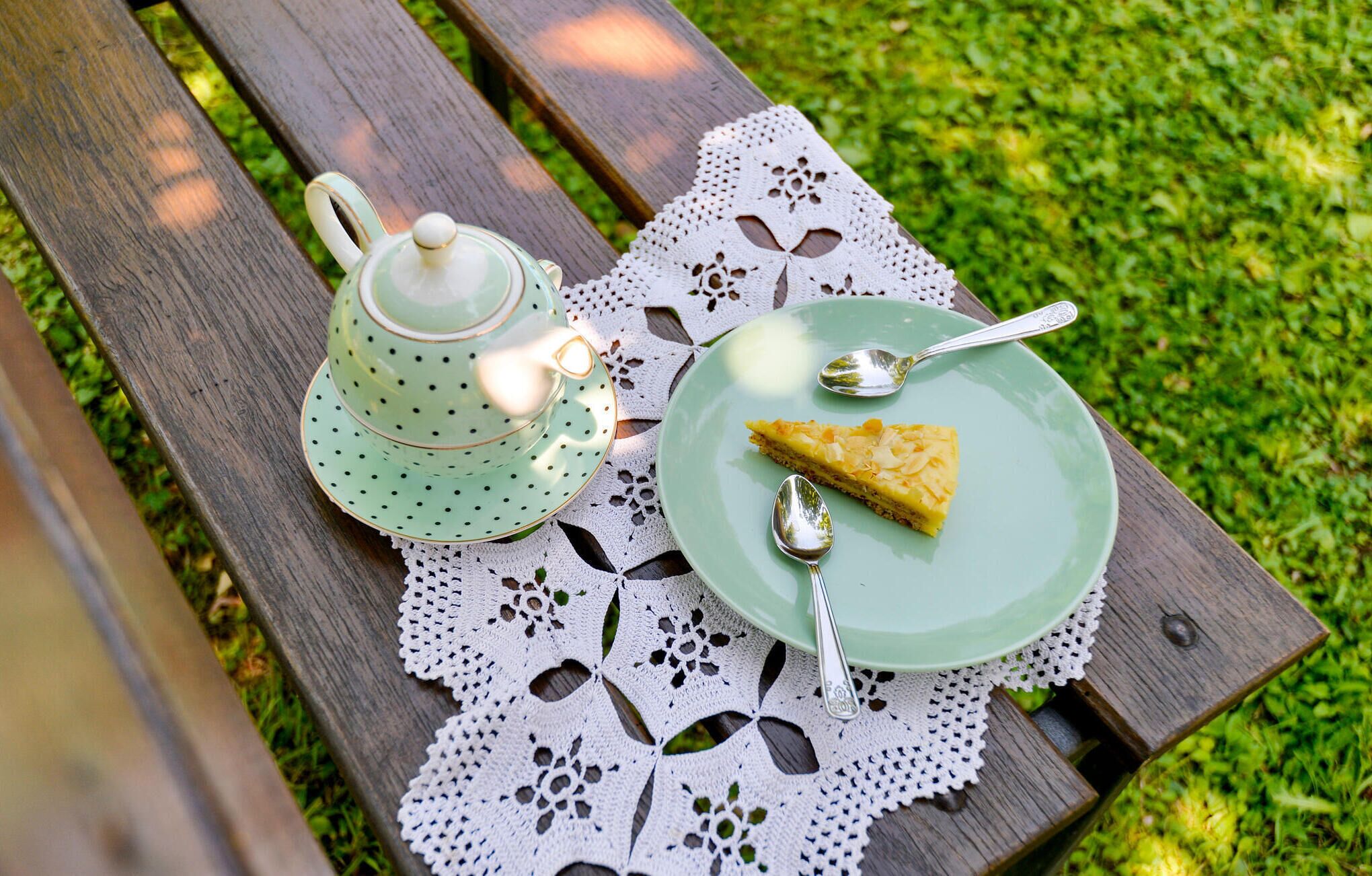 A picnic setup on a wooden bench with a lace tablecloth, a green teapot, a plate with a slice of pie, and two spoons.