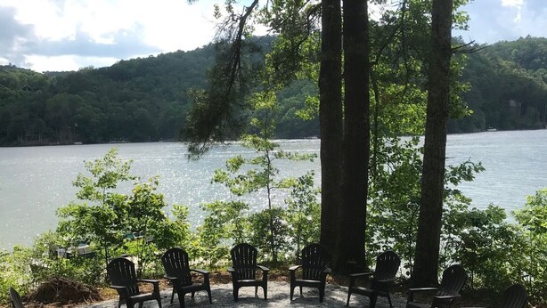 A lakeside seating area with Adirondack chairs and a fire pit, surrounded by trees and a view of the lake.