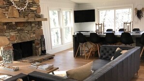 A modern living room with a stone fireplace, a large black sectional sofa, a dining area with a long table and chairs, and a flat-screen TV mounted on the wall.
