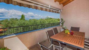 A balcony with a table set for two, a potted plant, and a view of greenery and a body of water. - Amazing home in Kozino (Kozino)