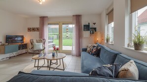 A living room with a sofa, a TV, a dining table, and a view of a garden through a glass door. - Awesome home in Wemeldinge with sauna (Wemeldinge)