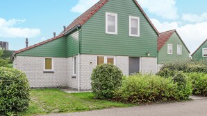 A two-story house with a green exterior and a red roof, featuring a stone foundation and a small front yard with bushes. - Awesome home in Wemeldinge with sauna (Wemeldinge)